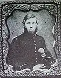 Framed portrait of a white man with a full bushy beard, wearing a dark military jacket and holding a tall feathered military hat.