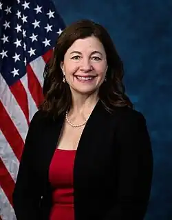 Official House portrait of McDonald Rivet smiling in front of the U.S. flag, wearing a black jacket, red shirt, and pearl necklace.