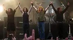 The members of Megadeth standing next to each other at a concert. They are holding their hands up.