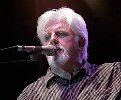 Man with silver shoulder-length hair and goatee in white v-neck top.