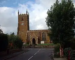 Red-brown church with limestone pinnacles and details.  A square tower on the left, the nave on the right under a low roof hidden behind castellations in the same material.  We can see a neat porch and a very imposing stained glass window.  The church and graveyard stand a metre higher than the road, retained behind a low wall of the same red-brown ironstone.  The church is seen framed by bushes and large trees in the middle nearground.  The sky is mostly of high, light, clouds with patches of blue.