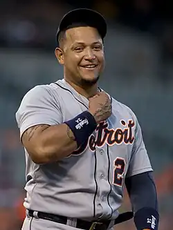 A brown-skinned man in a gray baseball jersey, with his right fist at the top of his chest.
