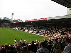 The inside of an association football stadium, with a stand on the right-hand side full of supporters. The pitch is visible to the left of the stand, with a floodlight in the background.