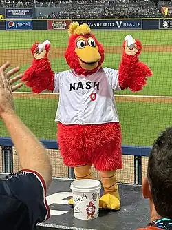A person wearing a red rooster costume dressed in a white baseball jersey with "NASH" on the chest in navy blue and a red "0" below it dances on a baseball dugout.