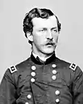 Head and shoulders of a young white man with wavy hair and a mustache, wearing a double-breasted military jacket with a rectangular patch over each shoulder.