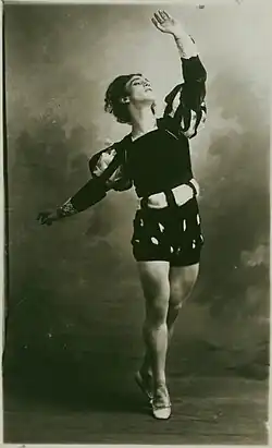 Black-and-white photograph of a costumed male dancer posed on half-toe, left arm raised upward, right arm extended backward.