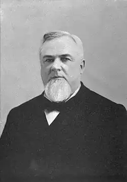 A moderately overweight man with a petite goatee and slicked back hair facing left wearing a black suit with a black bowtie