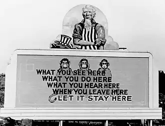 Uncle Sam has removed his hat and is rolling up his sleeves. On the wall in front of him are three monkeys and the slogan: What you see here / What you do here / What you hear here / When you leave here / Let it stay here.