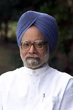A portrait photograph of a bespectacled Indian man with a dark grey beard, a blue turban, and a white button-down shirt standing before a natural backdrop of trees. A pen is in his pocket.