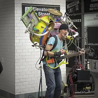 A man playing the guitar on a sidewalk. He has many musical instruments on him.