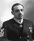 Head and torso of a man with a mustache and dark, thinning hair wearing a dark jacket with stripes on the upper sleeve, two round medals on the left breast, and a star-shaped medal hanging from a ribbon around his neck.