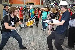 A man with a prop Half-Life 2 gravity gun (left) aiming his prop gun at another man with a prop Portal gun (right), at a convention with a crowd in the background.