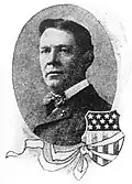 Head of a white man wearing a dark suit and bow tie. To the lower right of the oval-shaped portrait is an illustration of a shield covered with stars and stripes, like the American flag, and a ribbon tied around the shield.