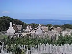A modern day photograph of a village consisting of small, primitive wooden houses.  Most of the houses have thatched roofs.  In the distance is a large expanse of ocean and a clear blue sky.  The village is surrounded by a wall consisting of tall, thick wooden planks.