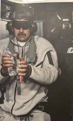 Photograph of Maduro standing blindfold, wearing earmuffs, and holding a plastic bottle of water