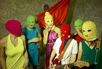 7 women with bright colored clothes and multicolored knit ski masks over their faces. A woman at the center holds a guitar and one at the back holds a piece of red fabric.