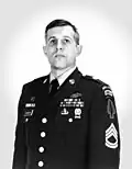 Head and torso of a white man standing erect and looking upwards, wearing a military jacket with an assortment of ribbon bars and badges on the left breast and chevrons and patches on the upper sleeve.