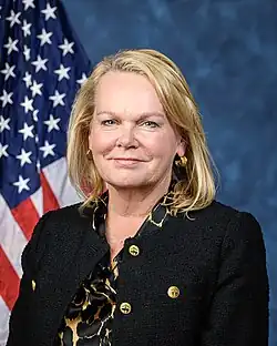 Official House portrait of McClain-Delaney smiling in front of the U.S. flag, wearing a black blazer jacket and a gold and black shirt.