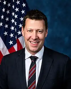 Official House portrait of Riley smiling in front of the U.S. flag, wearing a black suit, white shirt, and striped red, blue, and black tie.