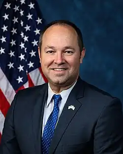 Official House portrait of Stutzman smiling in front of the U.S. flag, wearing a black suit with American and Israeli flags lapel pin, white shirt, and blue tie with white dots.