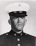 Head and shoulders of a young white man with a cleft chin wearing a dark military jacket and a white peaked cap with a black visor.