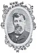 Head of a white man with bushy beard and thick hair wearing a suit, vest, and bow tie. The portrait is surrounded by a decorative oval frame.