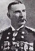Head and shoulders of a white man with a drooping mustache, wearing a military jacket with shoulder boards, one medal at the neck, and a row of medals across the entire chest.