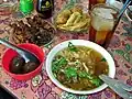 A selection of Indonesian food, including Soto Ayam (chicken soup), sate kerang (shellfish kebabs), telor pindang (preserved eggs), perkedel (fritter), and es teh manis (sweet iced tea)