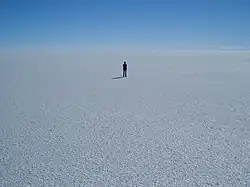 A panorama of flat, white ground, stretching to the horizon; a single person stands in the middle of the frame in the distance