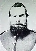 Head of a heavy-set white man with a beard and pointed mustache, wearing a military jacket.