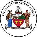 Circular seal with central images of a shield at center and sailing ship above it, with a European man to the left and a Native American to the right. The seal's edge reads "THE SEAL OF THE CITY OF ALBANY" with "ASSIDUITY" in a banner above the bottom.