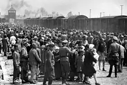 Large number of people standing beside a railway siding with the camp gate in the background