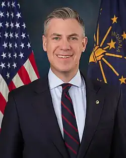Official Senate portrait of Banks smiling in front of the U.S. and Indiana flags, wearing a black suit with gold lapel pin, light blue shirt, and striped black and red tie.