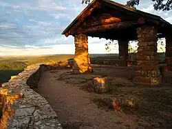A small, square fieldstone shelter with a roof at the end of a hiking trail. Looking over the edge of the fieldstone retaining wall is the dark green treetops of the Ozark National Forest as the sun sets behind a cloudy sky