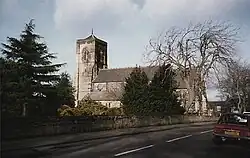 A church with a red tiled roof and a square tower. Trees in the foreground partially obscure the building. The sky is overcast and grey.