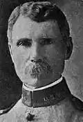 Head of a white man with wavy hair and a bushy mustache wearing a military jacket with the letters "U.S.V." on the high, stiff collar.