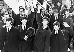 A black-and-white image of Paul McCartney, George Harrison, John Lennon and Ringo Starr waving to fans after arriving in America in 1964. A crowd is visible behind them on the left.