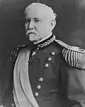 An older white man with Van Dyke mustache and beard wearing an ornate military jacket with fringe at the shoulders, a wide sash across the chest, and a medal hanging from his neck.