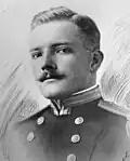 Head and shoulders of a white man with a neatly trimmed mustache wearing a military jacket with shoulder boards and two columns of buttons down the chest.