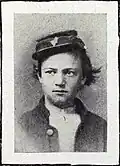Head and shoulders of a young white man with unkempt hair sticking out from under a tilted forage cap. He is wearing an open jacket over a white shirt.