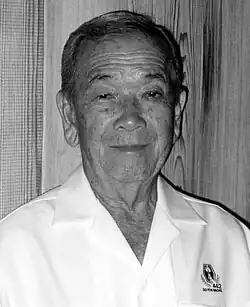 Head and shoulders of an elderly man wearing a white button shirt with an emblem on the left breast